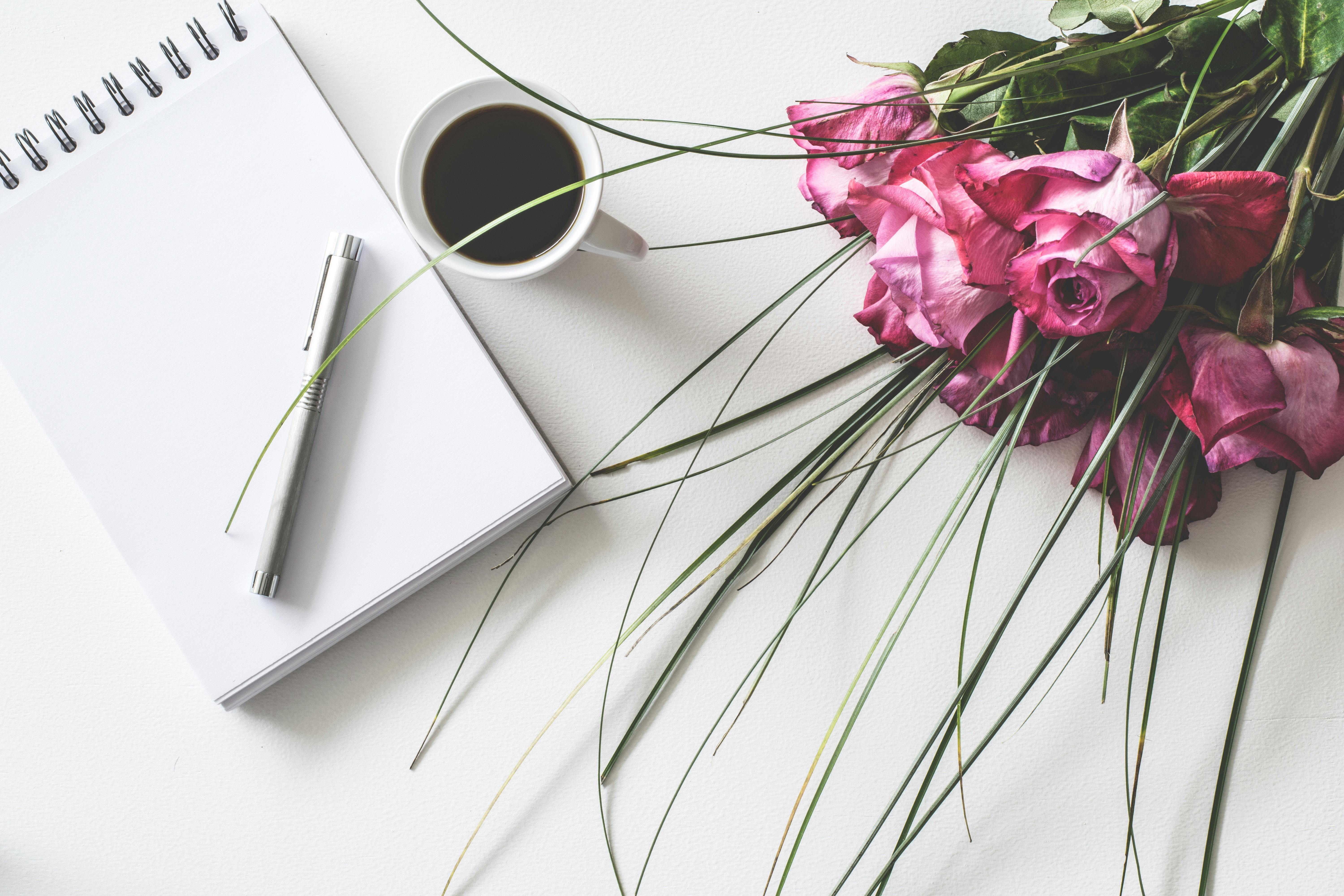 photograph of a notebook and pen, black coffee and pink roses